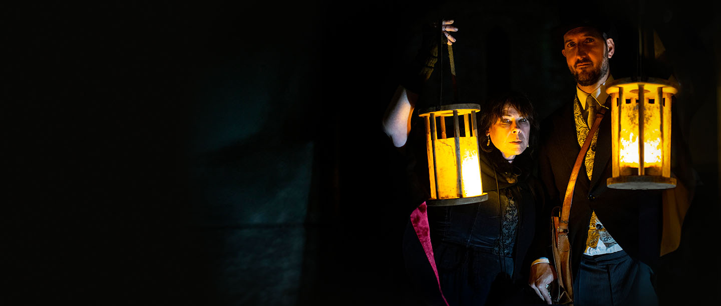 Image: Two people in Victorian dress standing in the dark with their faces lit by lanterns