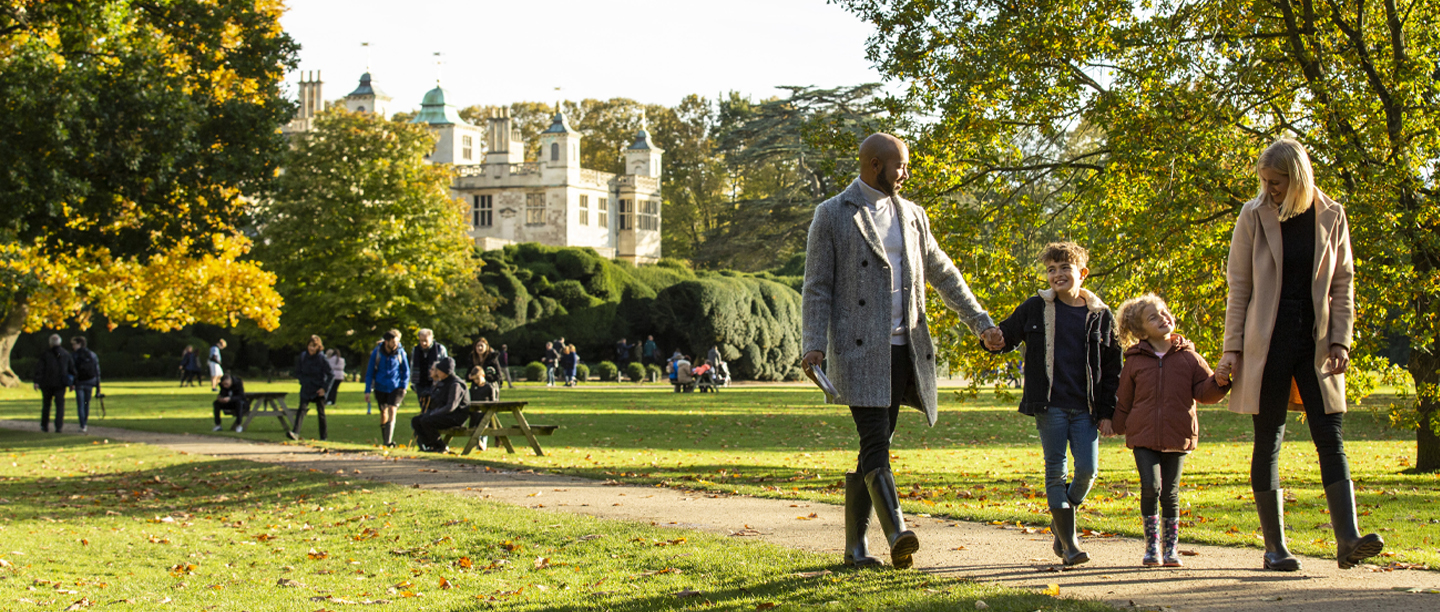 An image of a family walking through the grounds of a country house holding hands. The trees are loosing their leaves and there are many brown leaves on the ground.