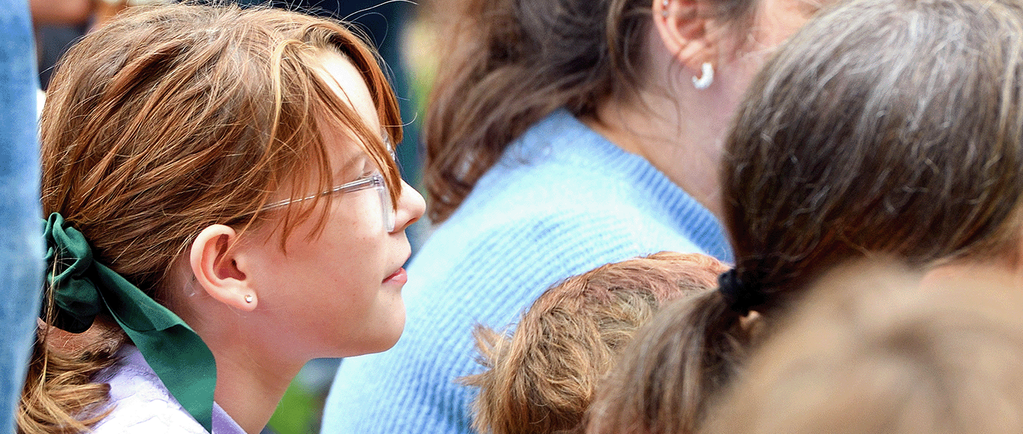 Photo of a young child with long hair and glasses watching an event in a crowd of people