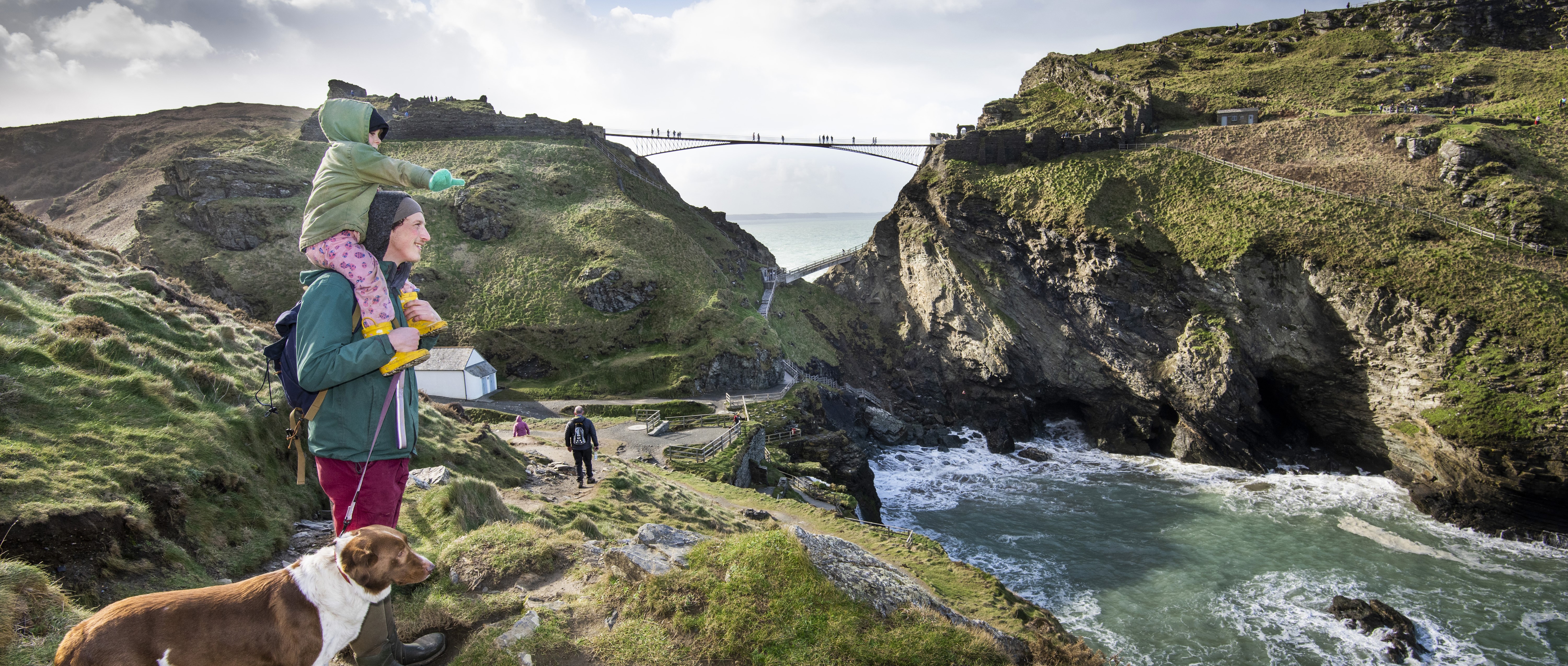 Photo of an adult with a child on their shoulders and a dog by their side looking out at the coast with the bridge at Tintagel Castle in the background